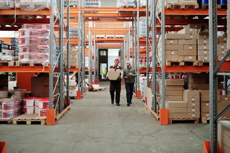 Two warehouse workers walking an aisle between tall racks of palletized goods